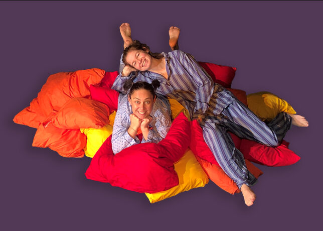 Zwei Frauen im Schlafanzug lächeln in die Kamera und liegen auf einem Kissenberg.