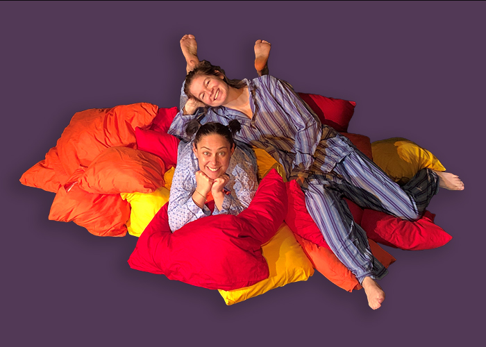 Zwei Frauen im Schlafanzug lächeln in die Kamera und liegen auf einem Kissenberg.